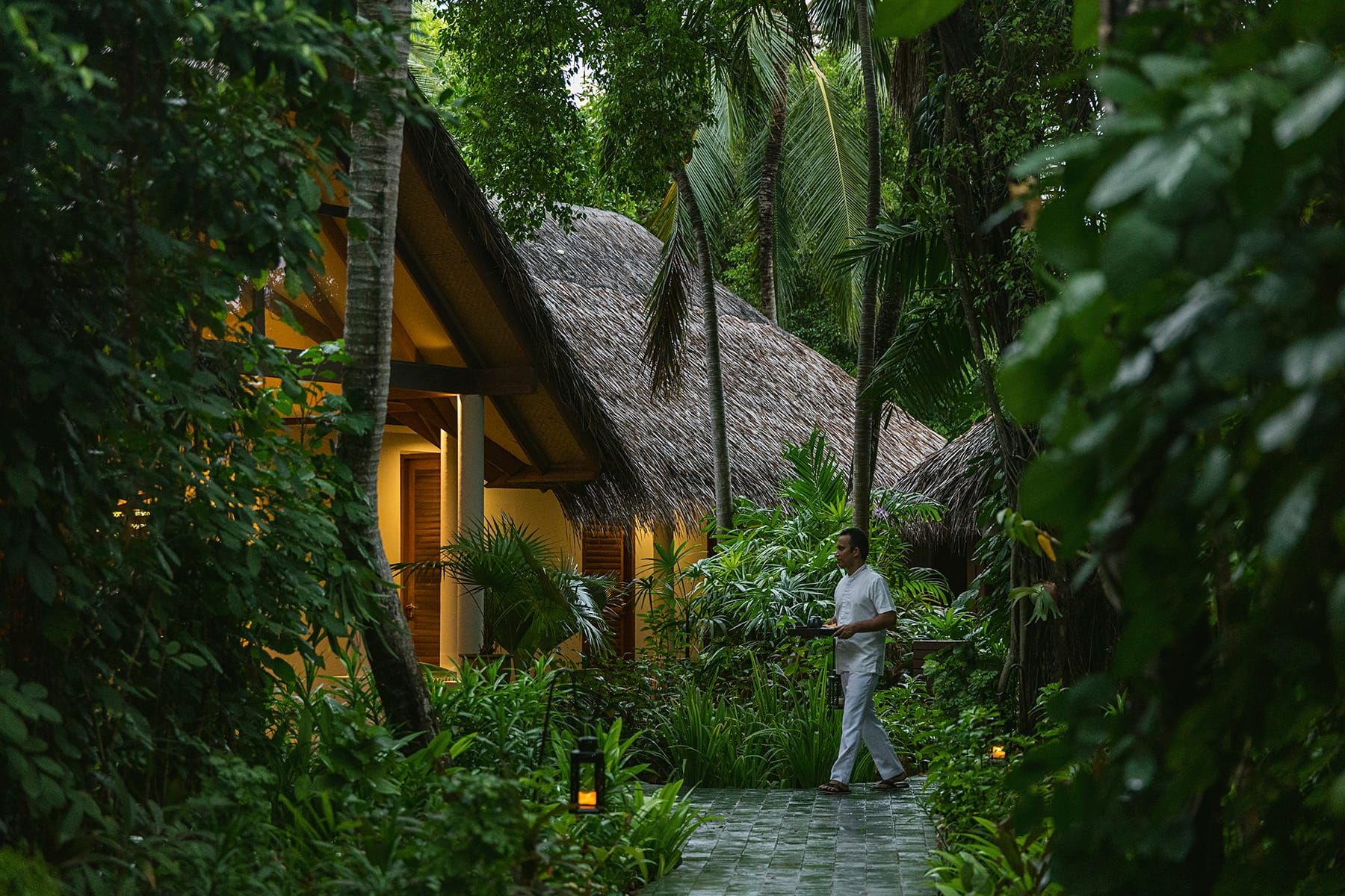 Therapist walking between thatched spa pavilions in a dense tropical garden at dusk