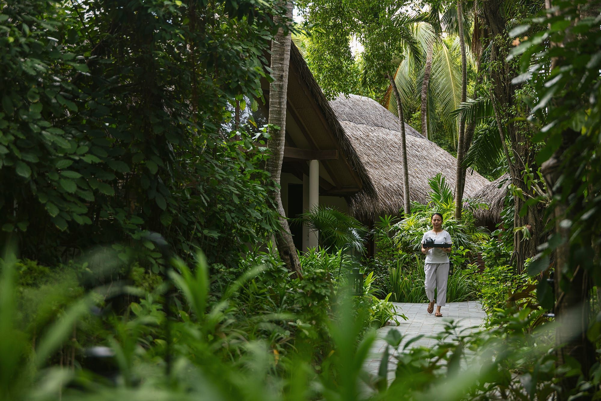 Therapist walking a stone path through lush jungle toward thatched spa pavilions
