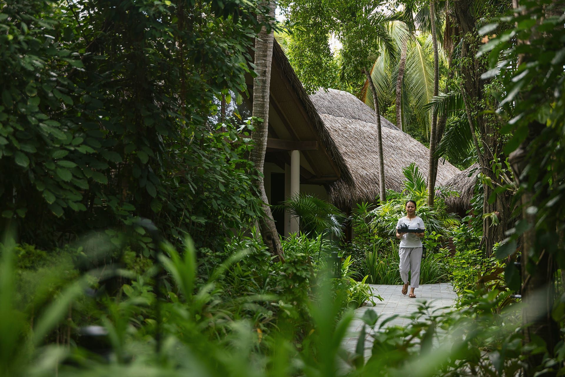Therapist walking a stone path through lush jungle toward thatched spa pavilions