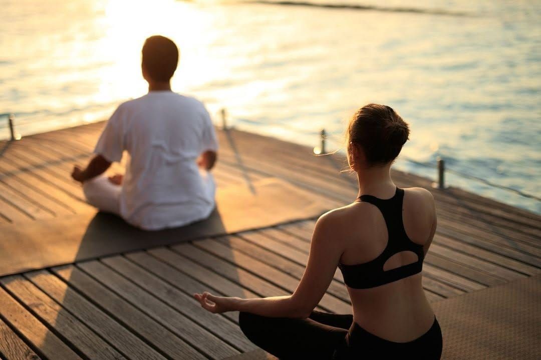Two people meditating cross-legged on a deck facing the sea at sunset